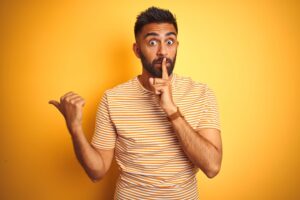 Man in yellow striped shirt with wide eyes holding one finger over his lips and pointing over his shoulder with his thumb